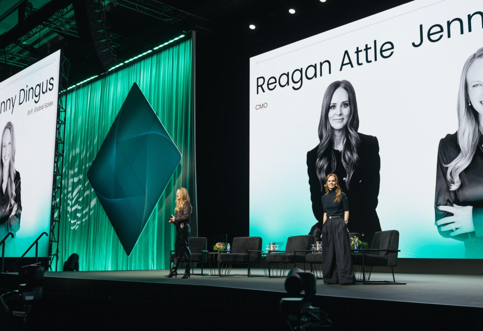 Two women stand on a stage with large screens displaying their portraits, under green lighting.