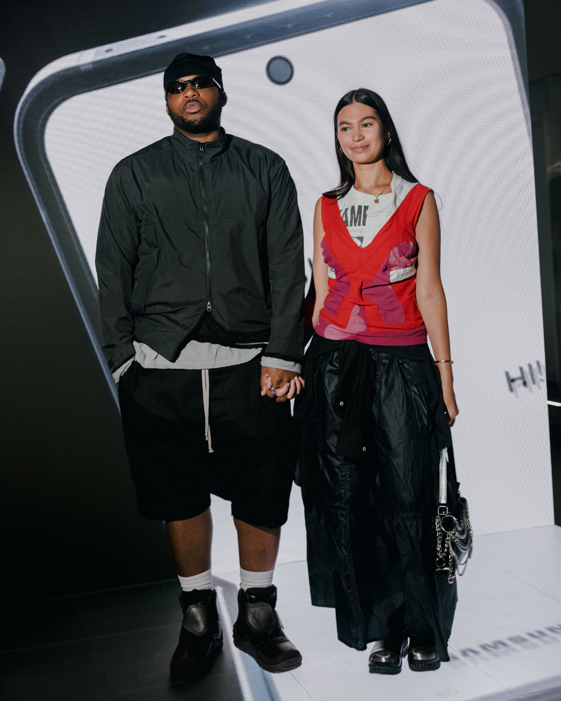 A man and woman are standing hand in hand, posing in front of a smartphone display.