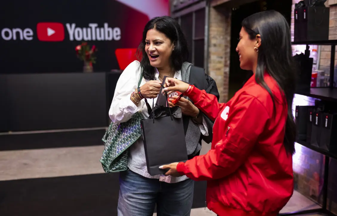 Deux femmes souriant et échangeant un sac cadeau noir, avec un logo YouTube en arrière-plan.