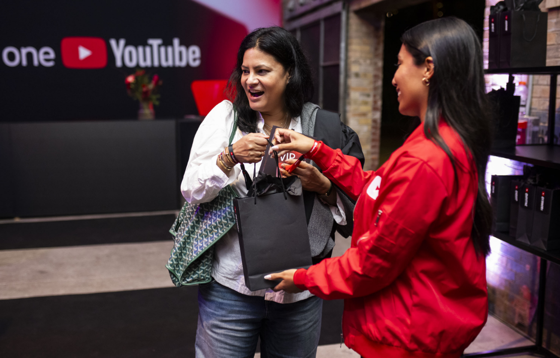 Two women smiling and exchanging a black gift bag, with a YouTube logo in the background.