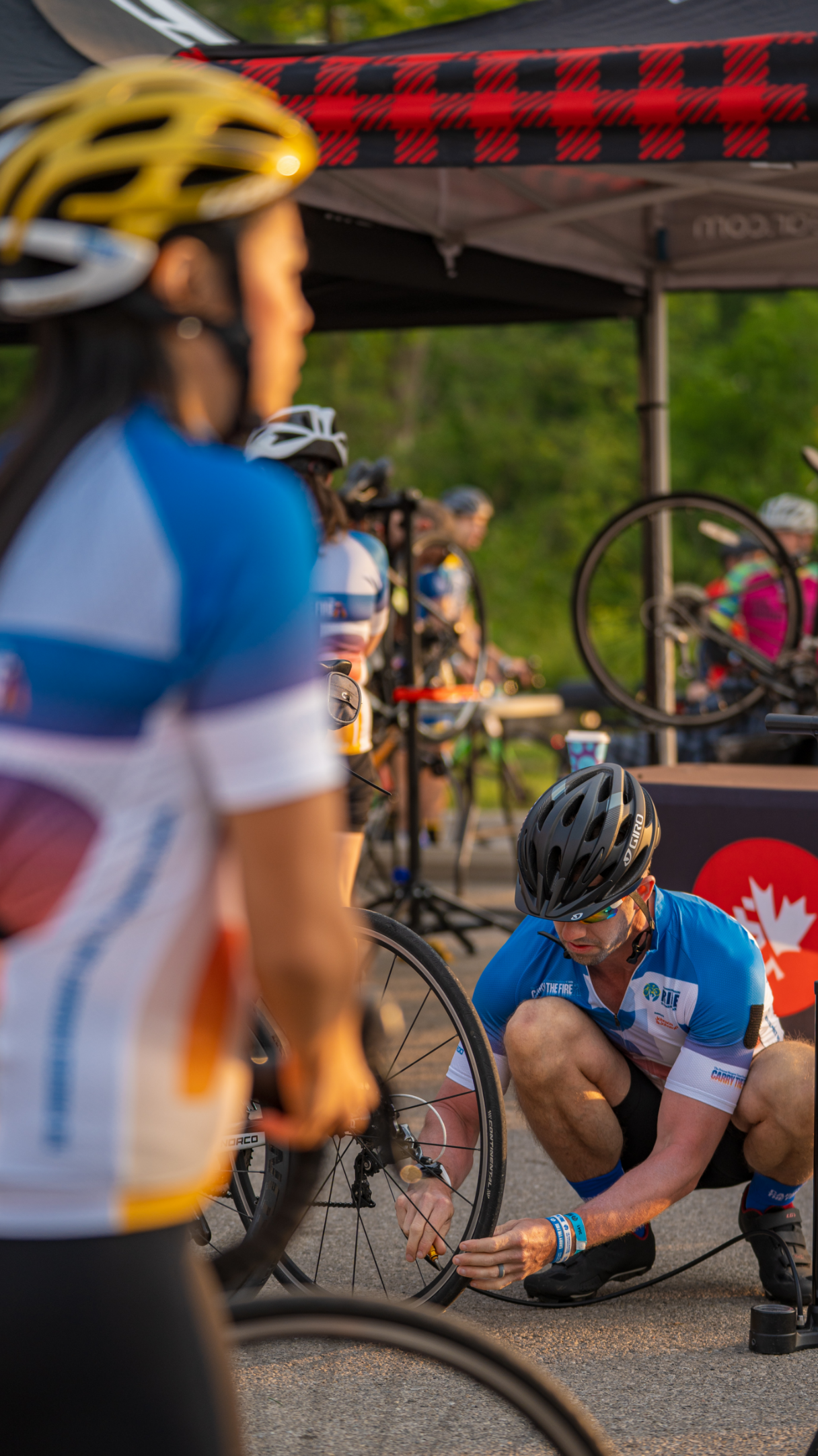 A group of cyclists in helmets and jerseys gather near a tent, with one person fixing a bike tire.