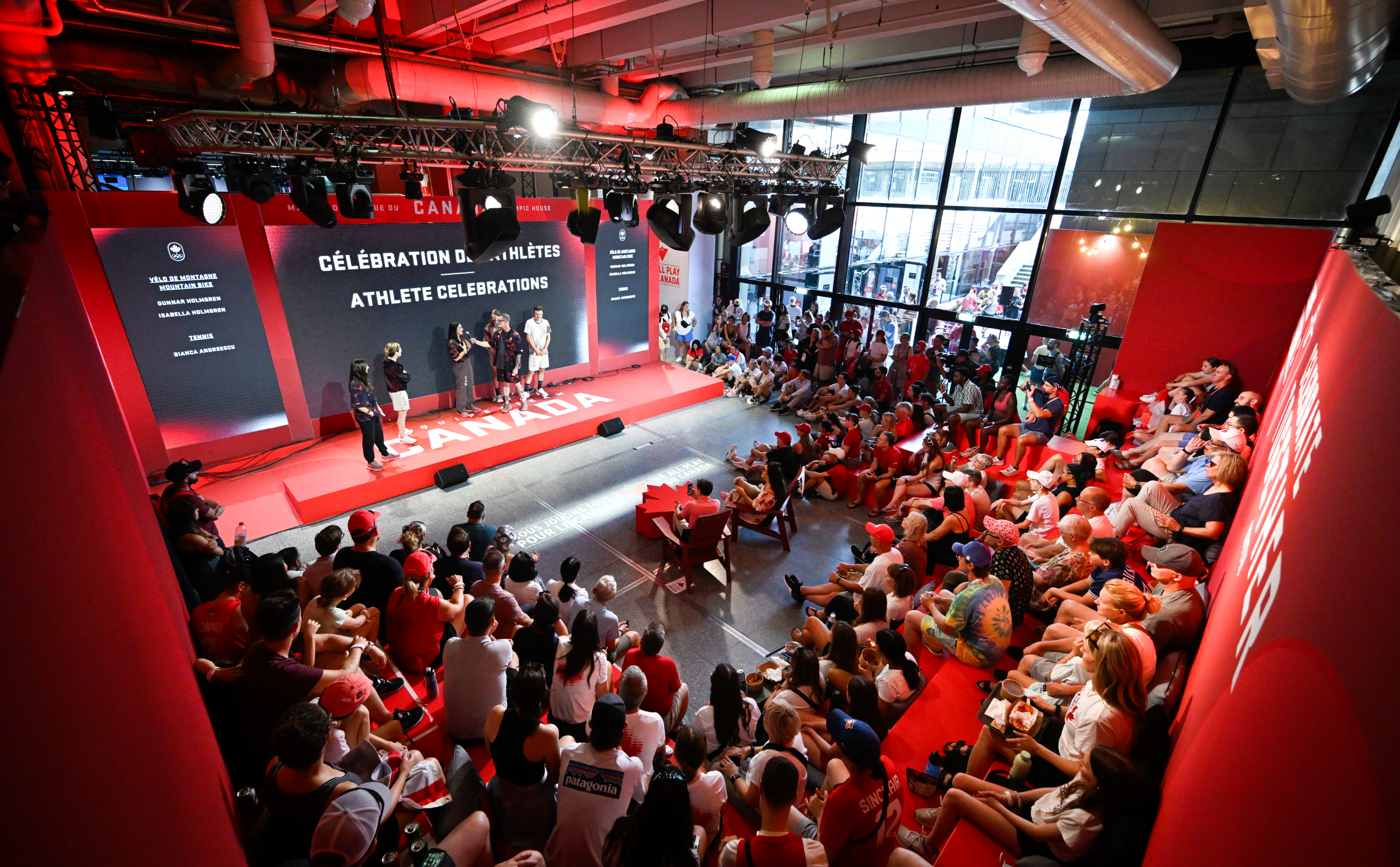 A large crowd gathers in a red-themed room watching a presentation on stage celebrating athletes.
