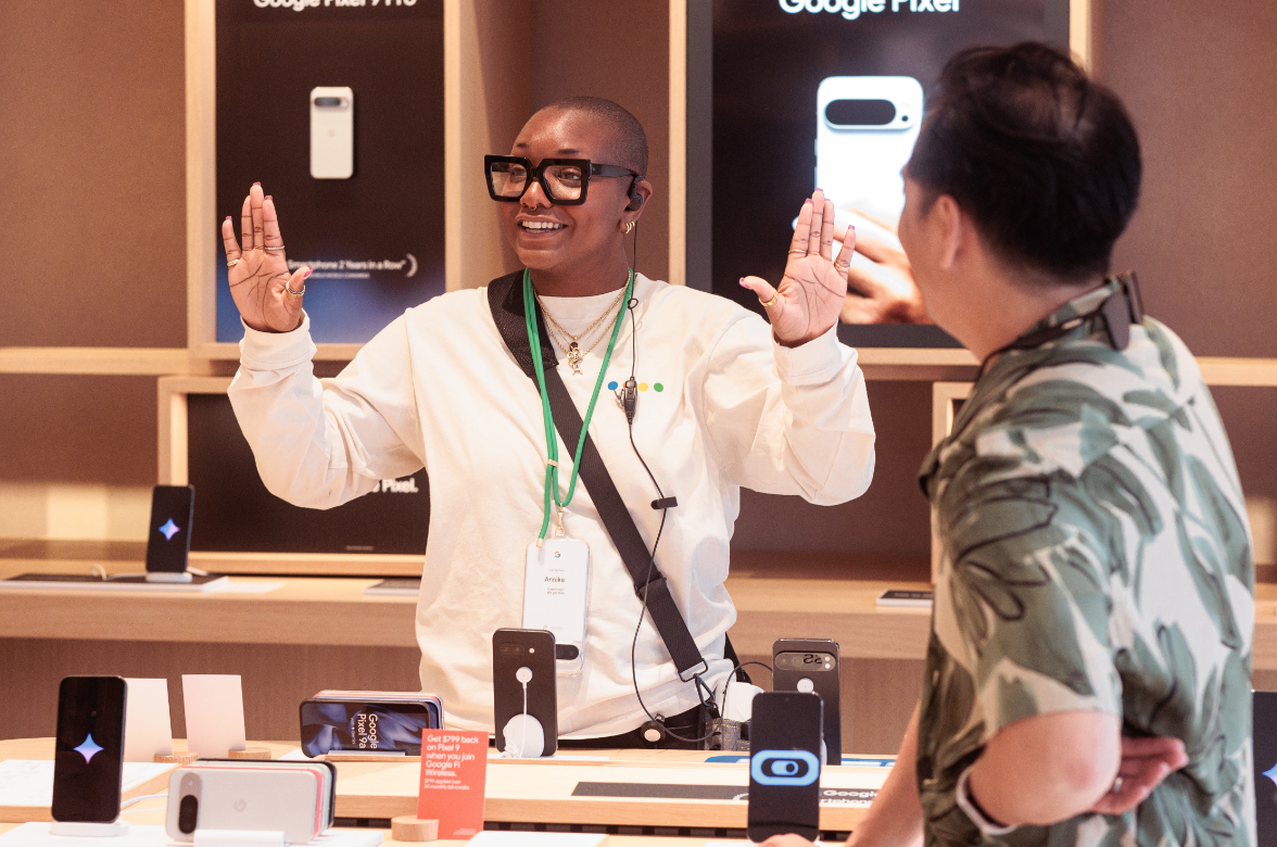 A person wearing a white shirt and glasses is enthusiastically speaking to another person in a store displaying Google Pixel devices.