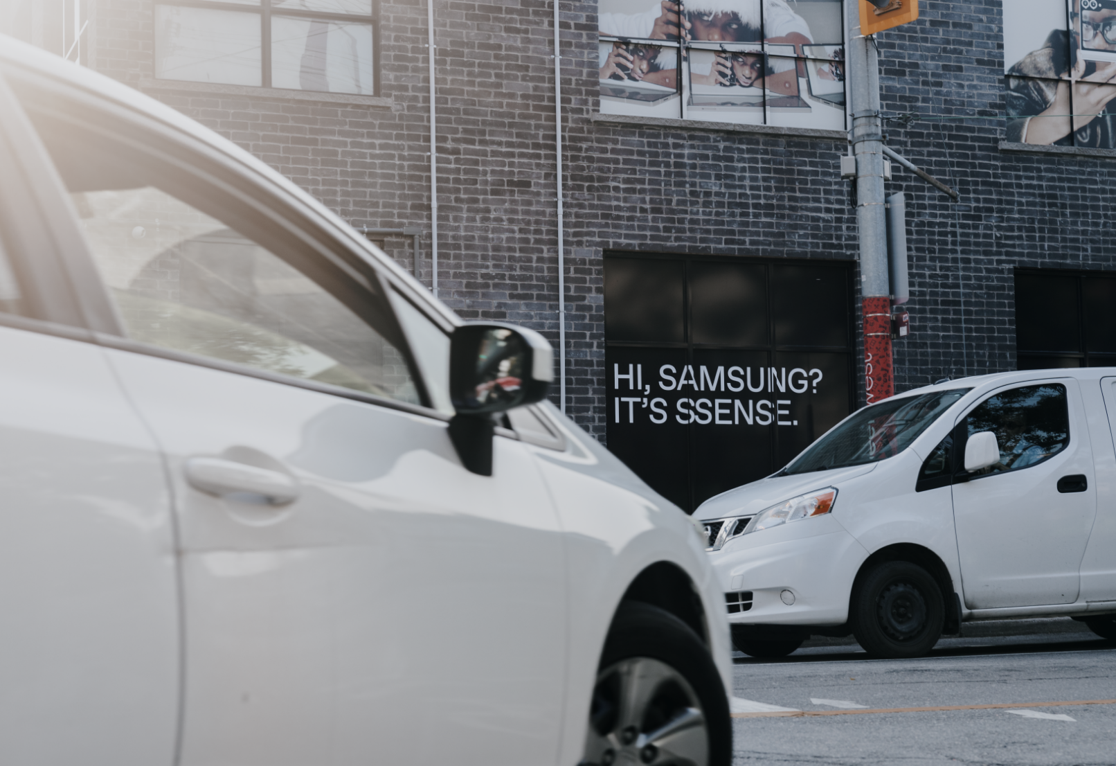 Cars parked on a street in front of a brick building with a message saying "Hi, Samsung? It's SSENSE."