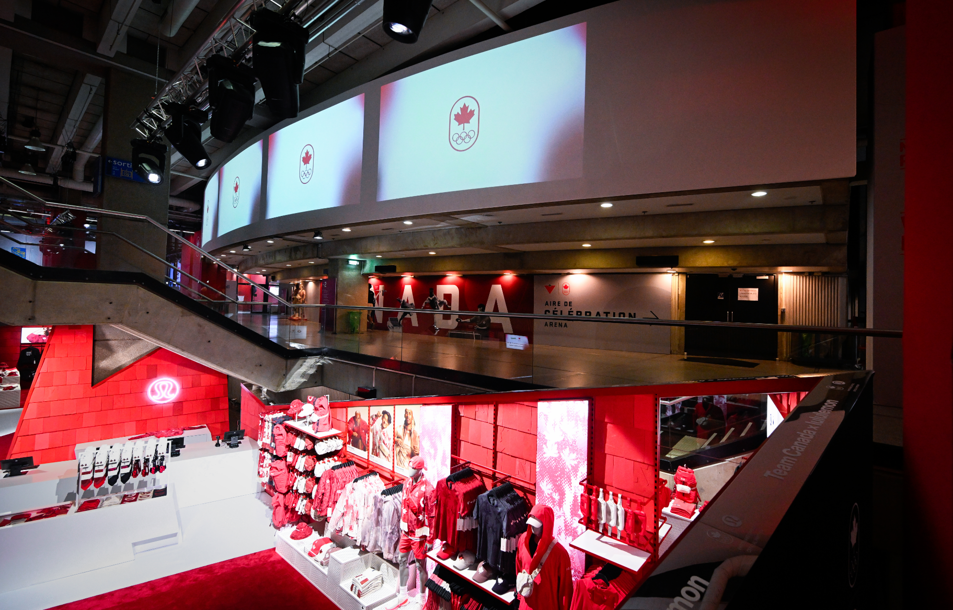A retail store with red merchandise displays beneath large screens showing a Canadian Olympic symbol.