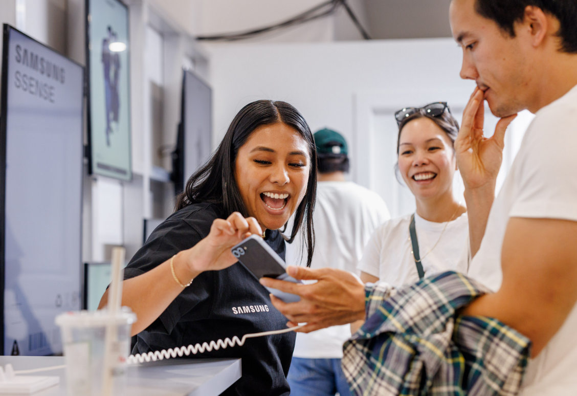 A Samsung employee shows a phone to two smiling customers in a store.