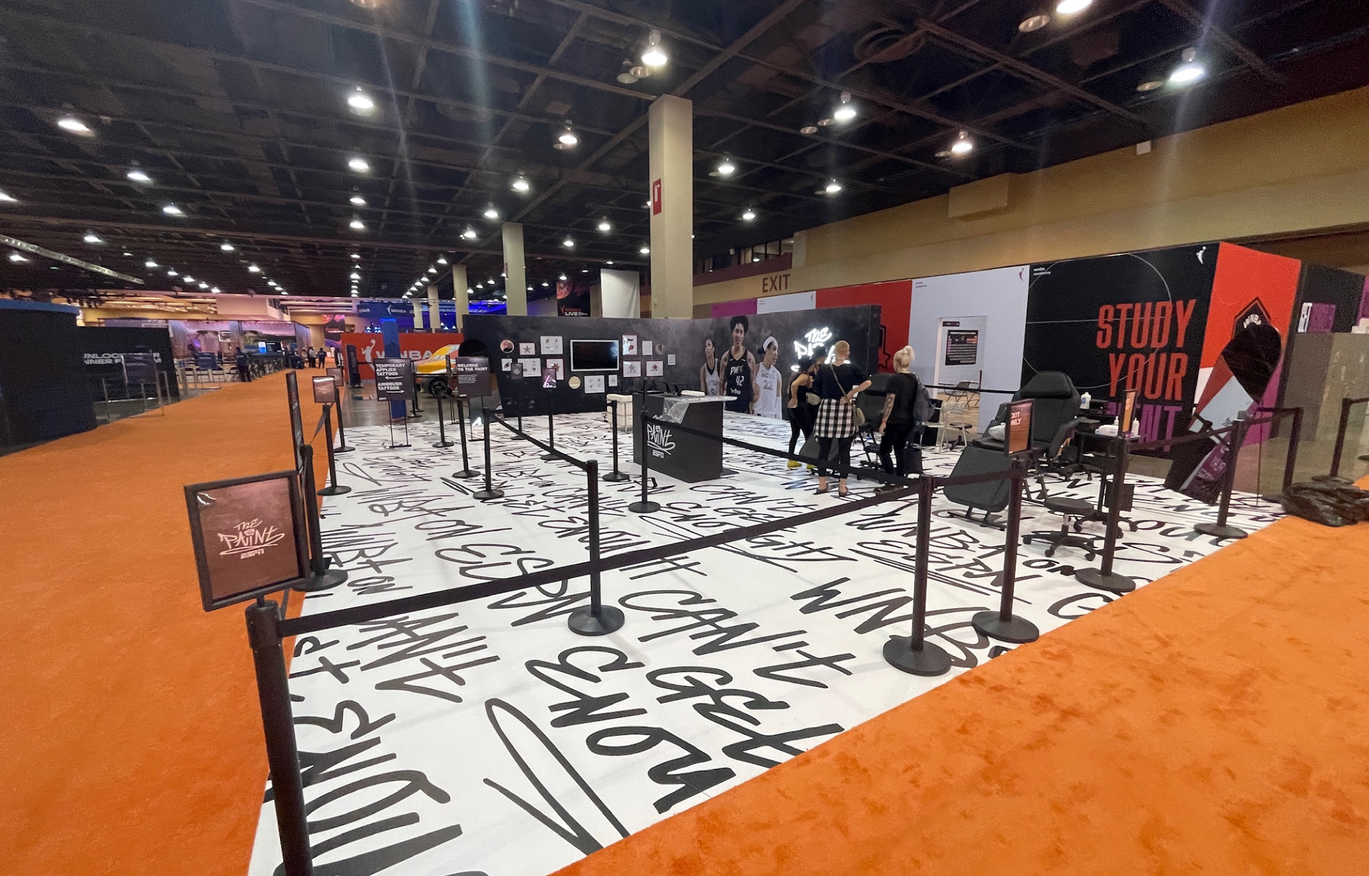 Spacious convention center area with art installations, black and white floor print, and people interacting at a display booth.
