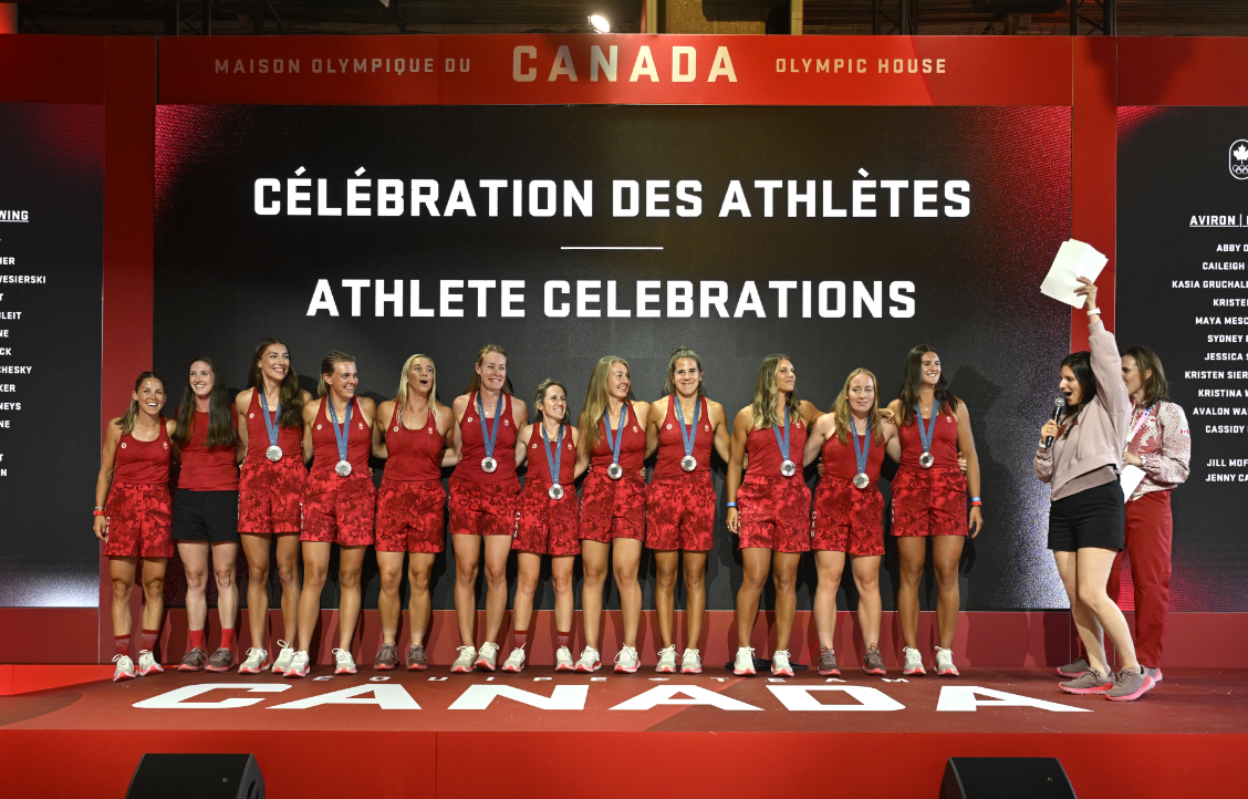A group of athletes wearing medals stands on a stage under a sign that reads "Athlete Celebrations" in both French and English.