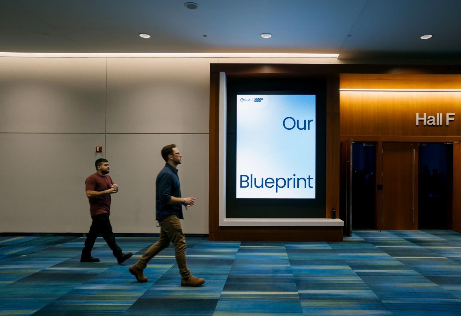 Two people walking in a hallway with a digital sign reading "Our Blueprint" and doors to Hall F.