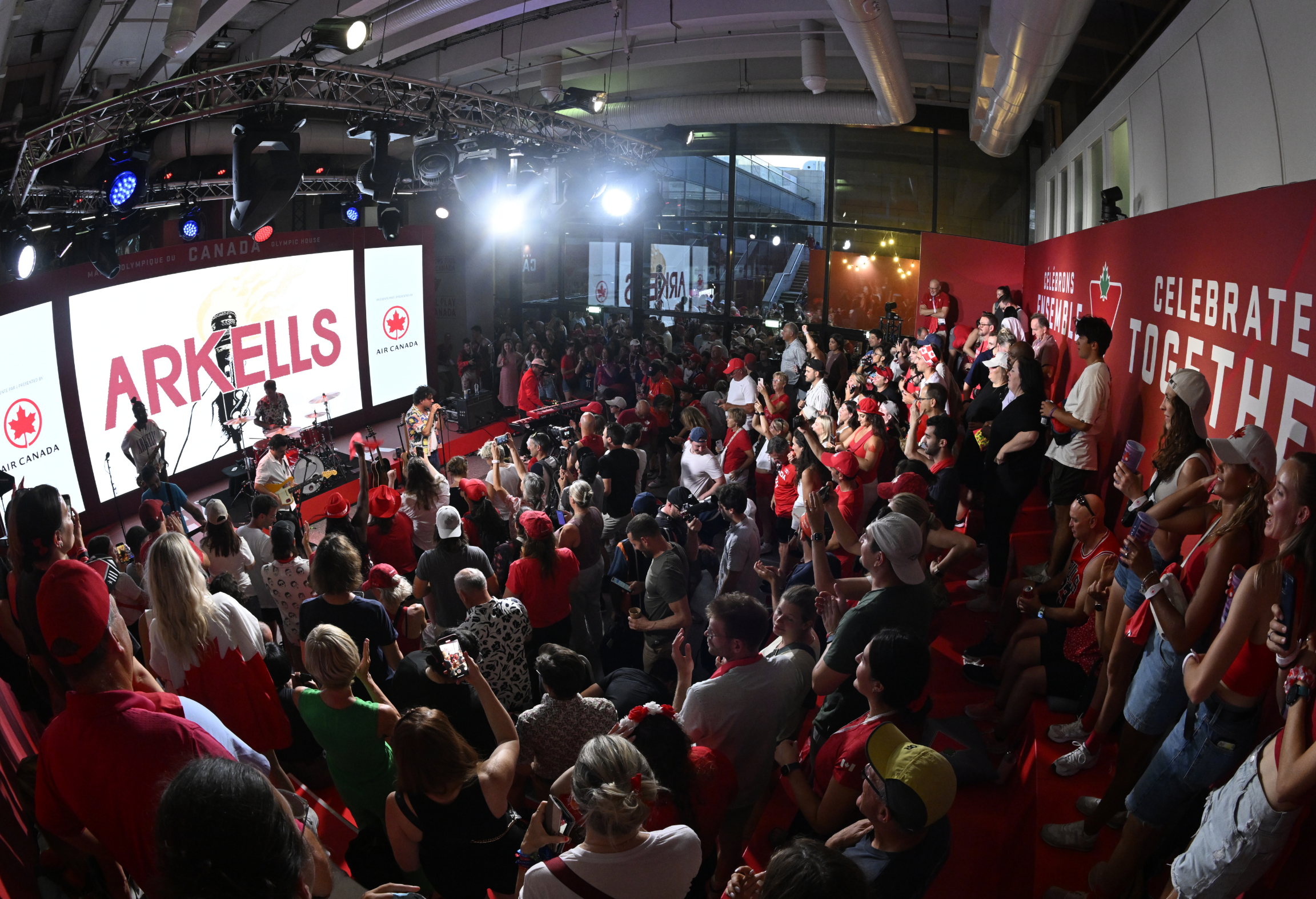 A band performs on stage in front of a large, cheering crowd inside a venue.