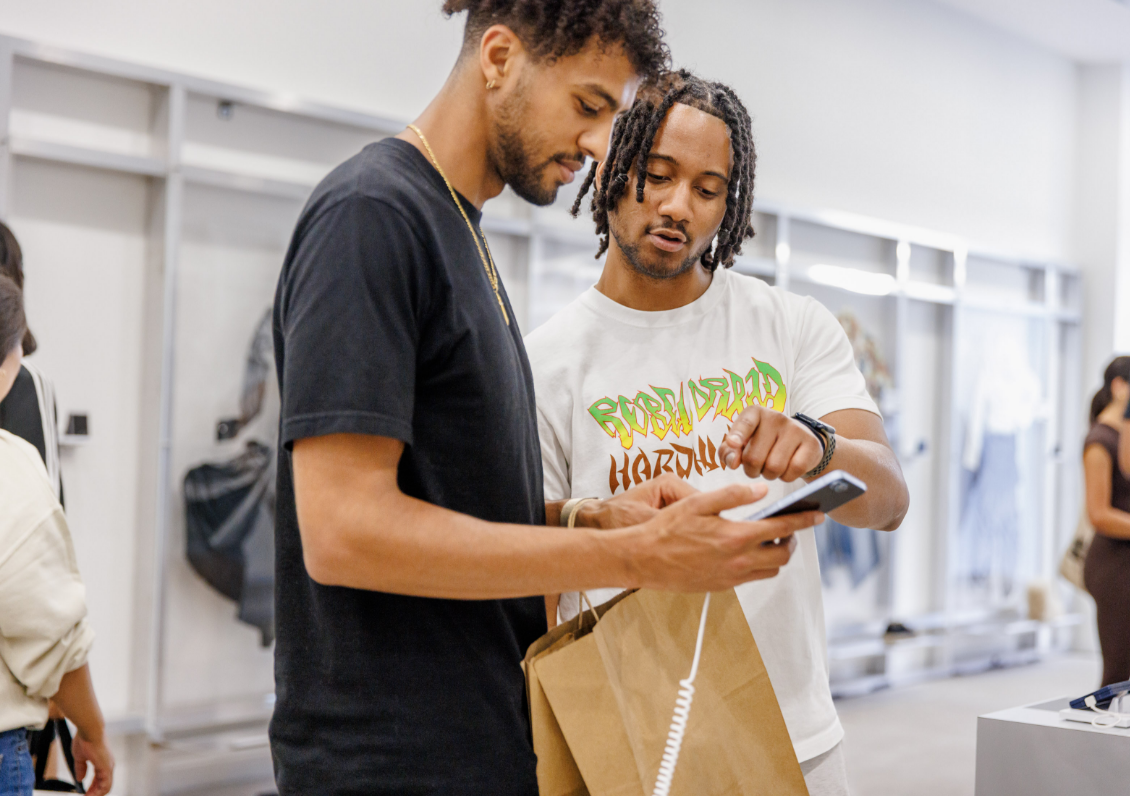 Two men are looking at a phone together in a store, one holding a shopping bag.