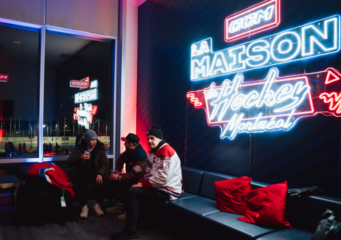 Three people sitting and talking on a couch beside a large neon sign that says "La Maison Hockey Montréal."