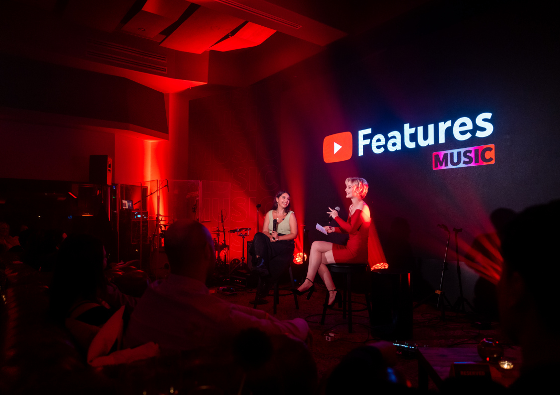 Two people are sitting on stools on a stage, having a conversation in front of a large screen with "Features Music" displayed.