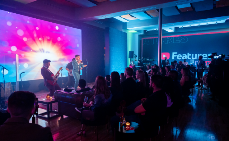 A live band performing on a stage with colorful lights in front of an audience at an indoor venue.