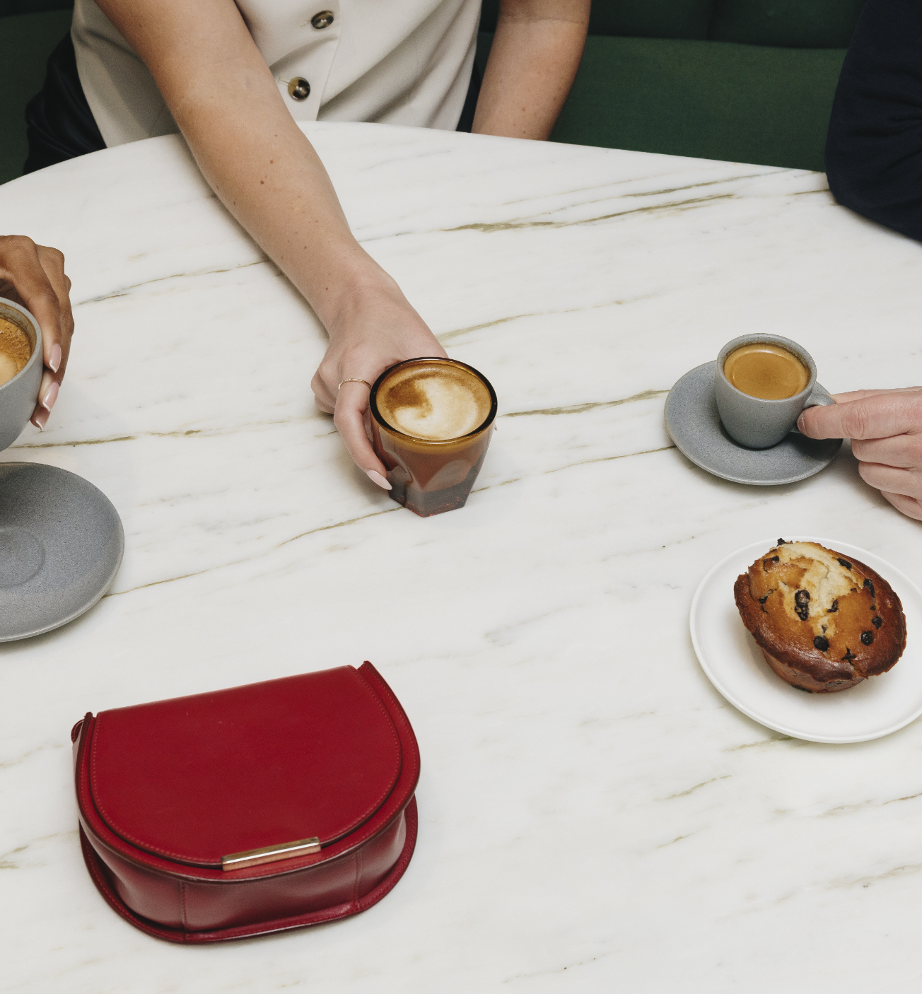 Three people enjoying coffee and a muffin at a marble table with a red purse placed nearby.
