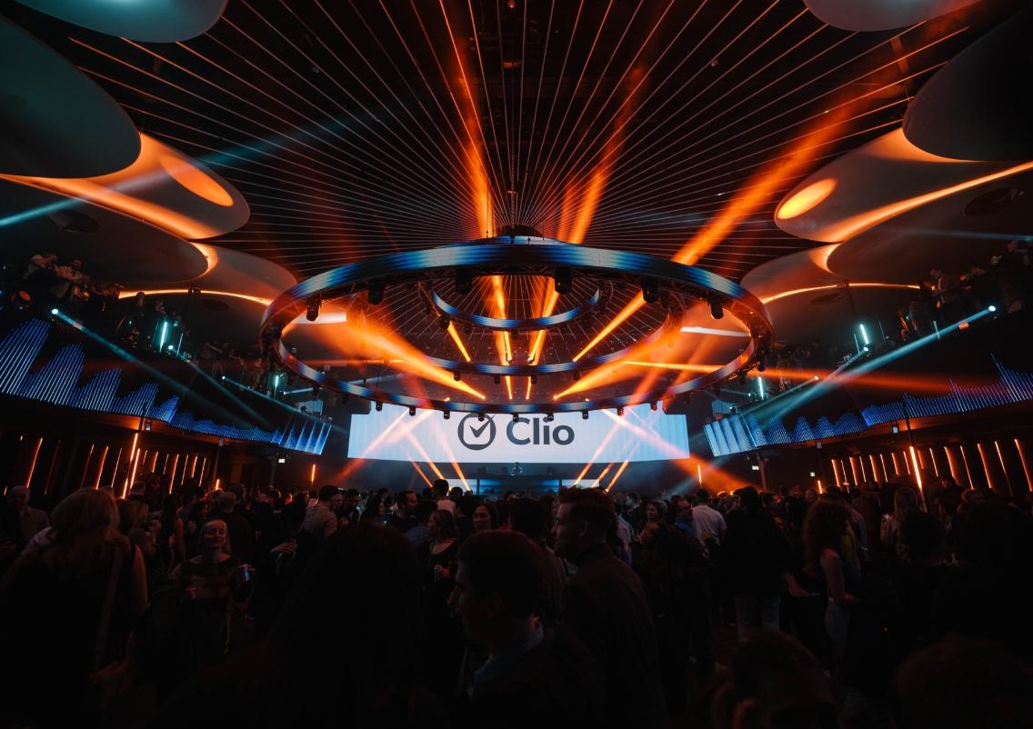 A lively nightclub scene with a crowd, colorful lighting, and a large screen displaying the name "Clio."