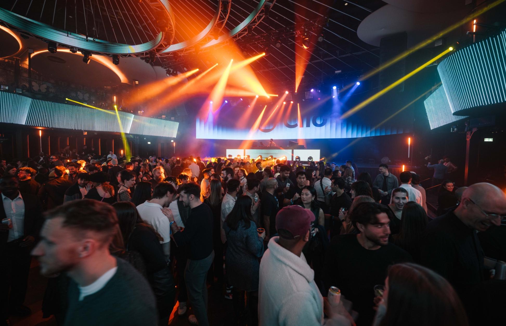 A crowded nightclub with colorful lights and a DJ playing music.