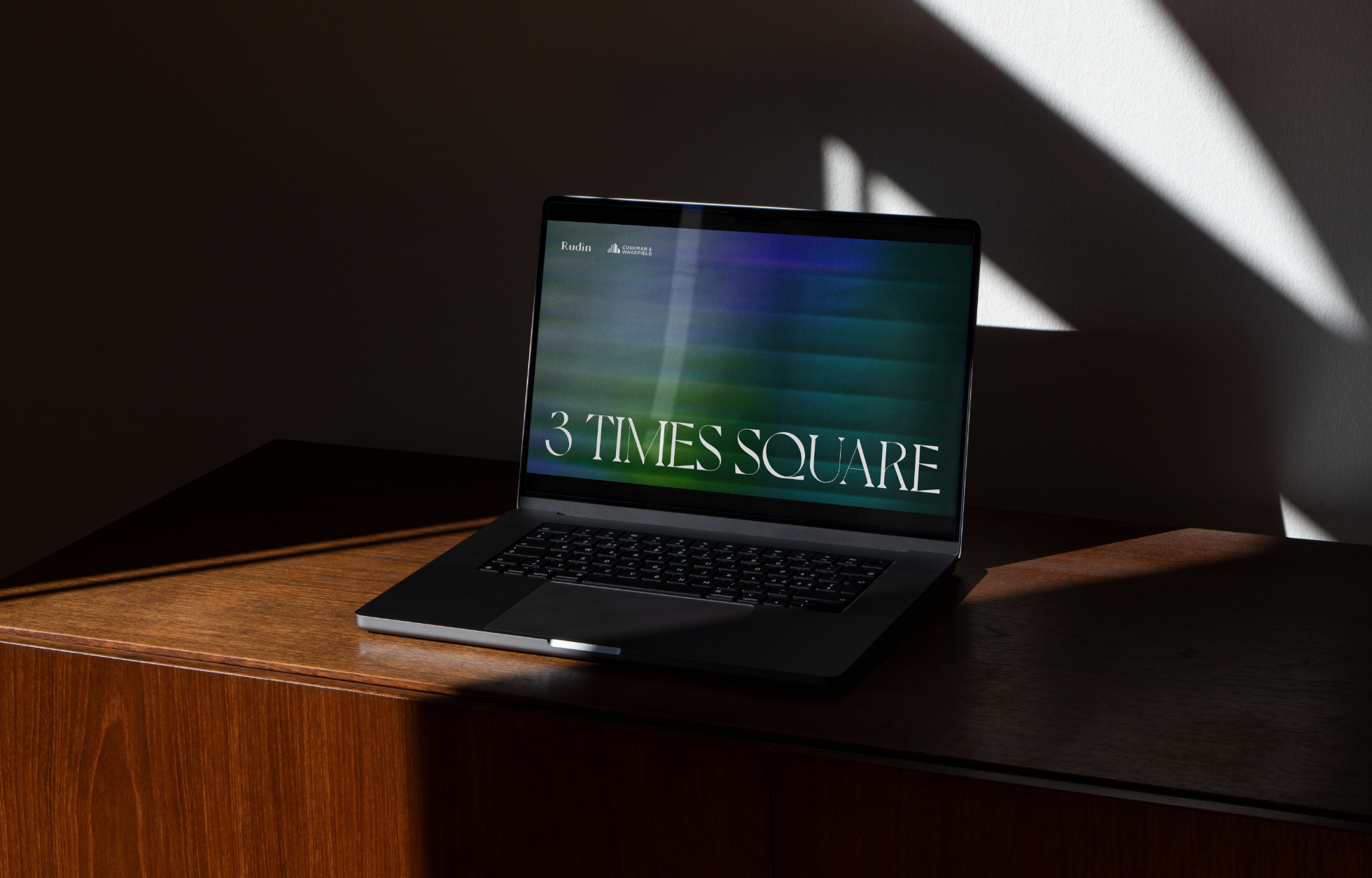 A laptop displaying "3 Times Square" on its screen is sitting on a wooden surface with dramatic shadow lighting.