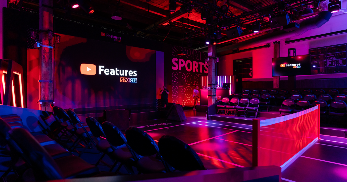 A dimly lit event space with a stage displaying "Features Sports" on a large screen, surrounded by rows of seats.