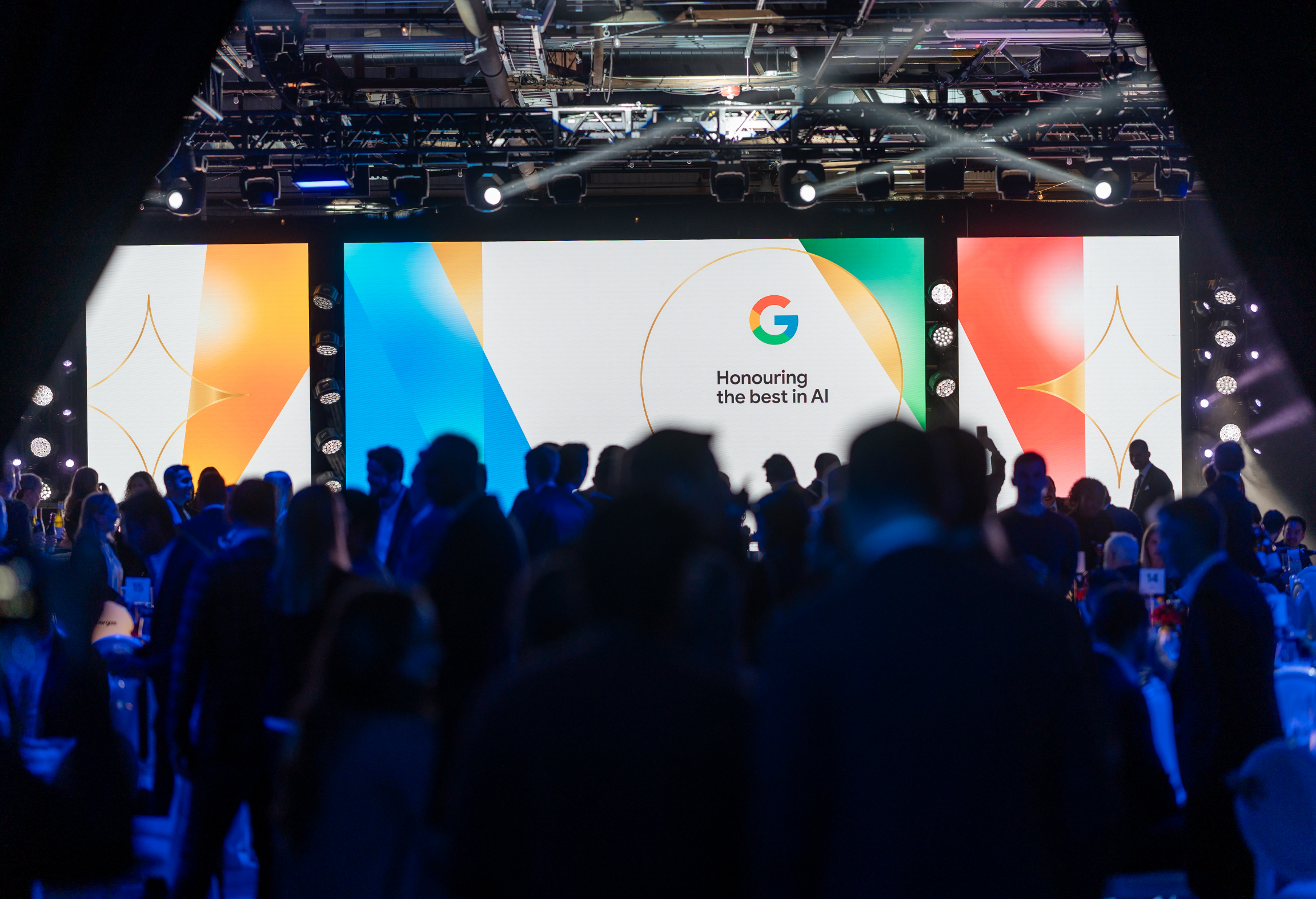 A crowd of people at an event with a large screen displaying "Honouring the best in AI."