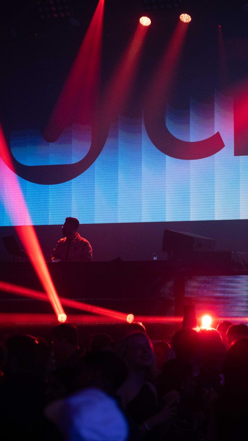 A DJ performing on stage with colorful lights and a crowd dancing in the foreground.