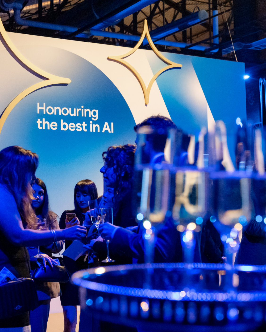 A group of people socializing at an event celebrating achievements in AI, with champagne glasses in the foreground and a sign that reads "Honouring the best in AI."
