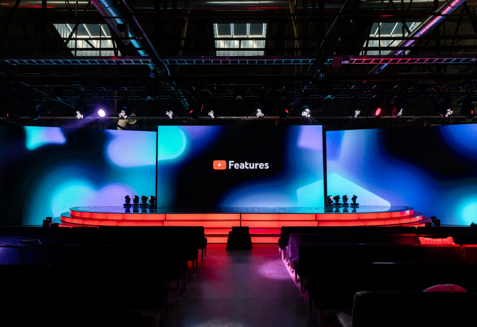 The image shows a stage setup with a large screen displaying the YouTube logo and the word "Features," surrounded by colorful lights and empty seating.