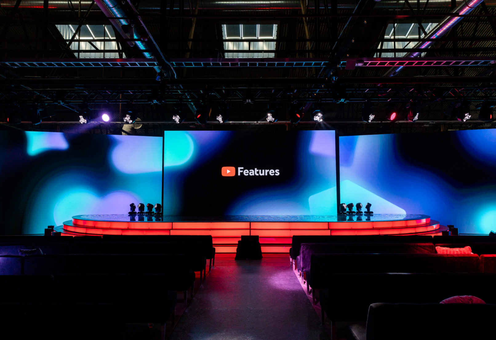 The image shows a stage setup with a large screen displaying the YouTube logo and the word "Features," surrounded by colorful lights and empty seating.