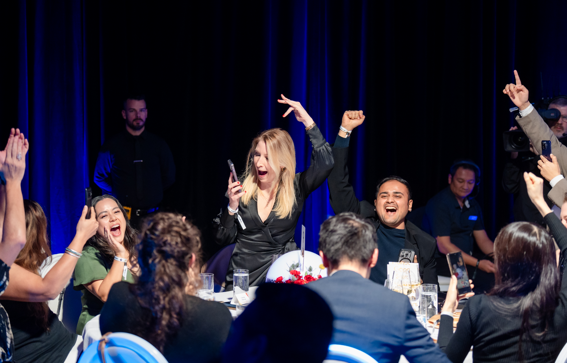 People at a table cheer and celebrate, some holding their phones up.