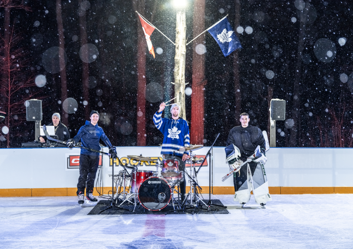 Trois personnes debout sur une patinoire de hockey extérieure dans la neige, l'une jouant de la batterie.