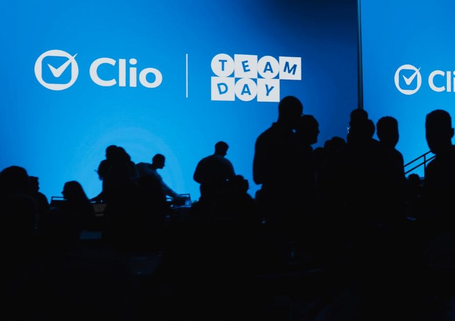 Silhouetted crowd in front of a blue screen displaying "Clio Team Day."