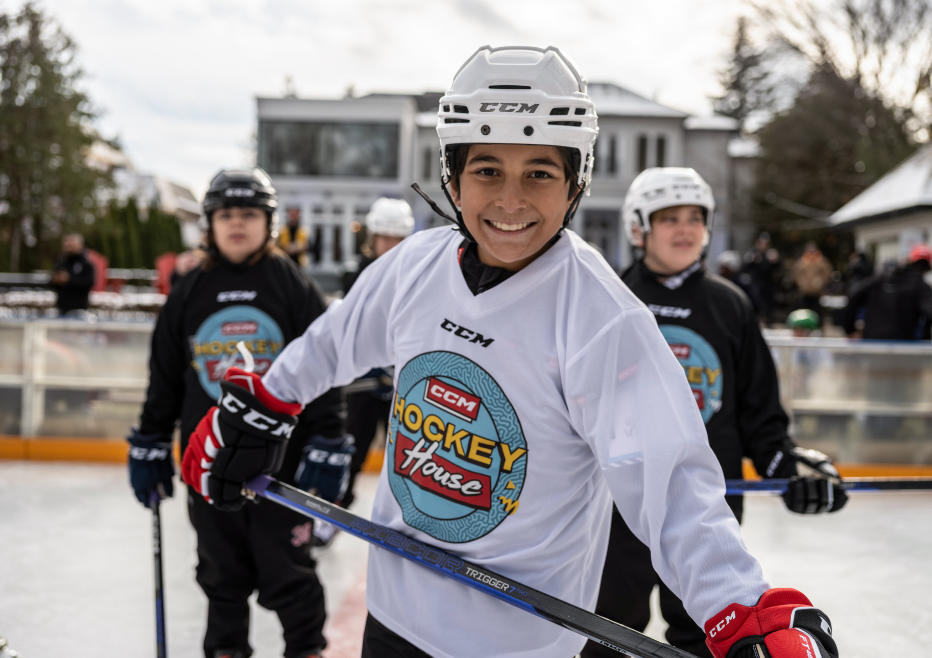 A young hockey player in a white jersey smiles on an outdoor ice rink with teammates in the background.