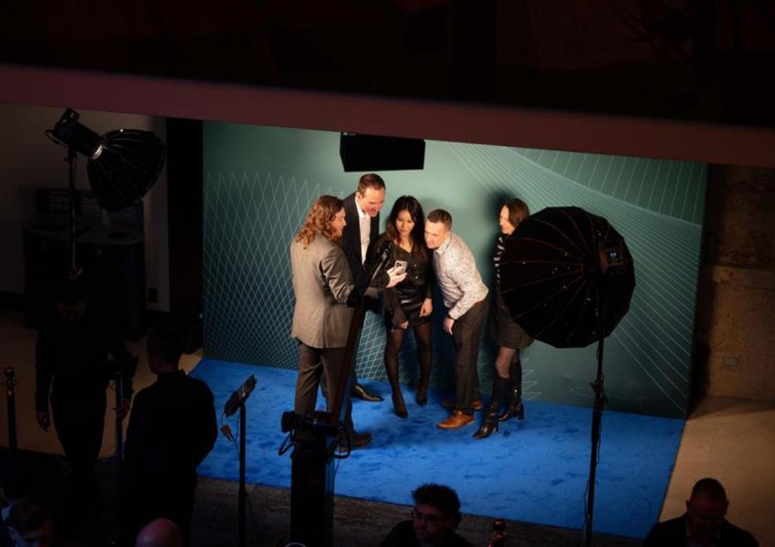 A group of five people are gathered around a smartphone under professional lighting on a blue carpet.
